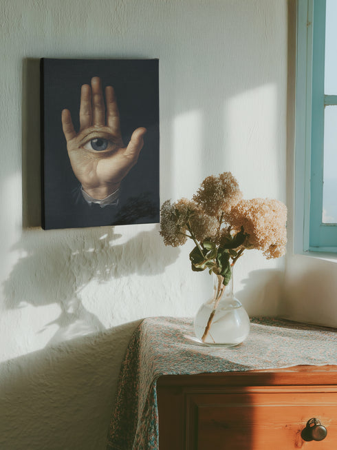 A high-contrast matte canvas print featuring a surrealist artwork. Centered against a dark, textured charcoal background, a realistic, pale hand is held up, palm forward. Embedded in the center of the palm is a detailed, light blue human eye, looking directly at the viewer. The hand emerges from a formal, dark suit cuff with a white ruffled sleeve, adding a touch of vintage, academic style. The overall mood is mysterious, gothic, and scholarly, suitable for dark academia or maximalist decor.